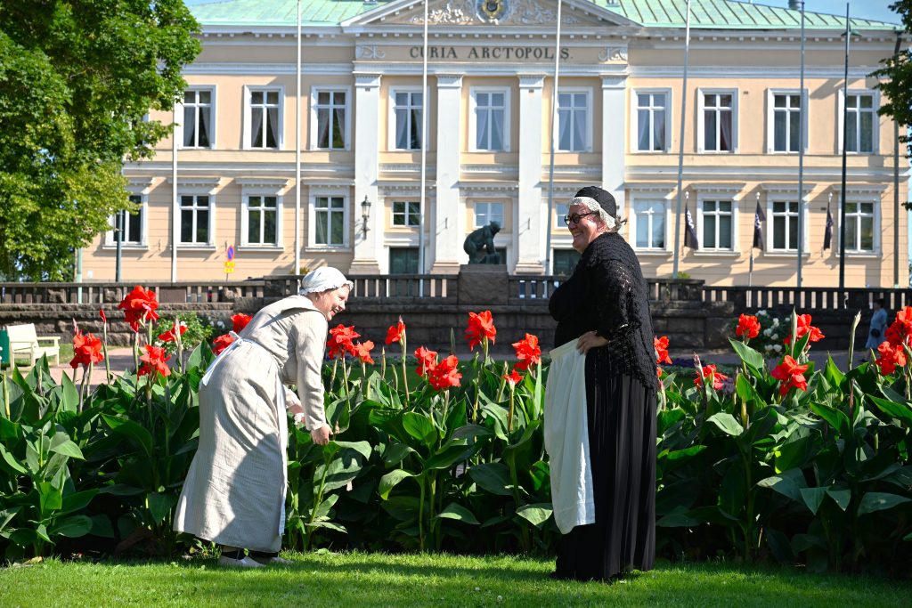 Vanhantyylisiin vaatteisiin pukeutuneet naiset raatihuoneen edustan puistomaisemassa.