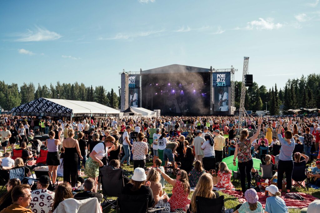 Auringonpaisteisena festivaalipäivänä paljon ihmisiä etualalla kuvassa ja ihmismassan takana näkyy esiintymislava