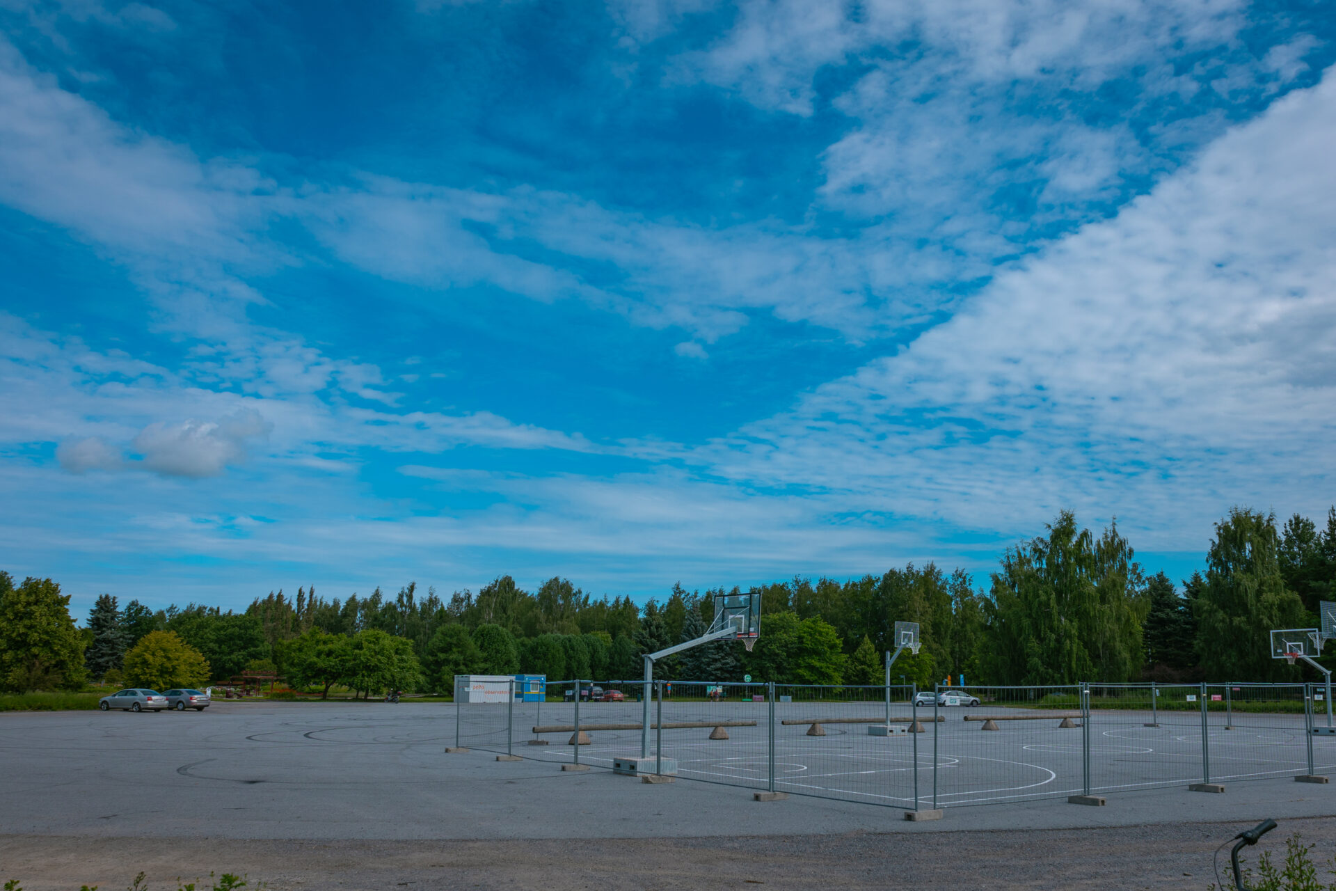 parkkipaikalla aidattu alue jossa koripallokenttä, taustalla autoja ja puita