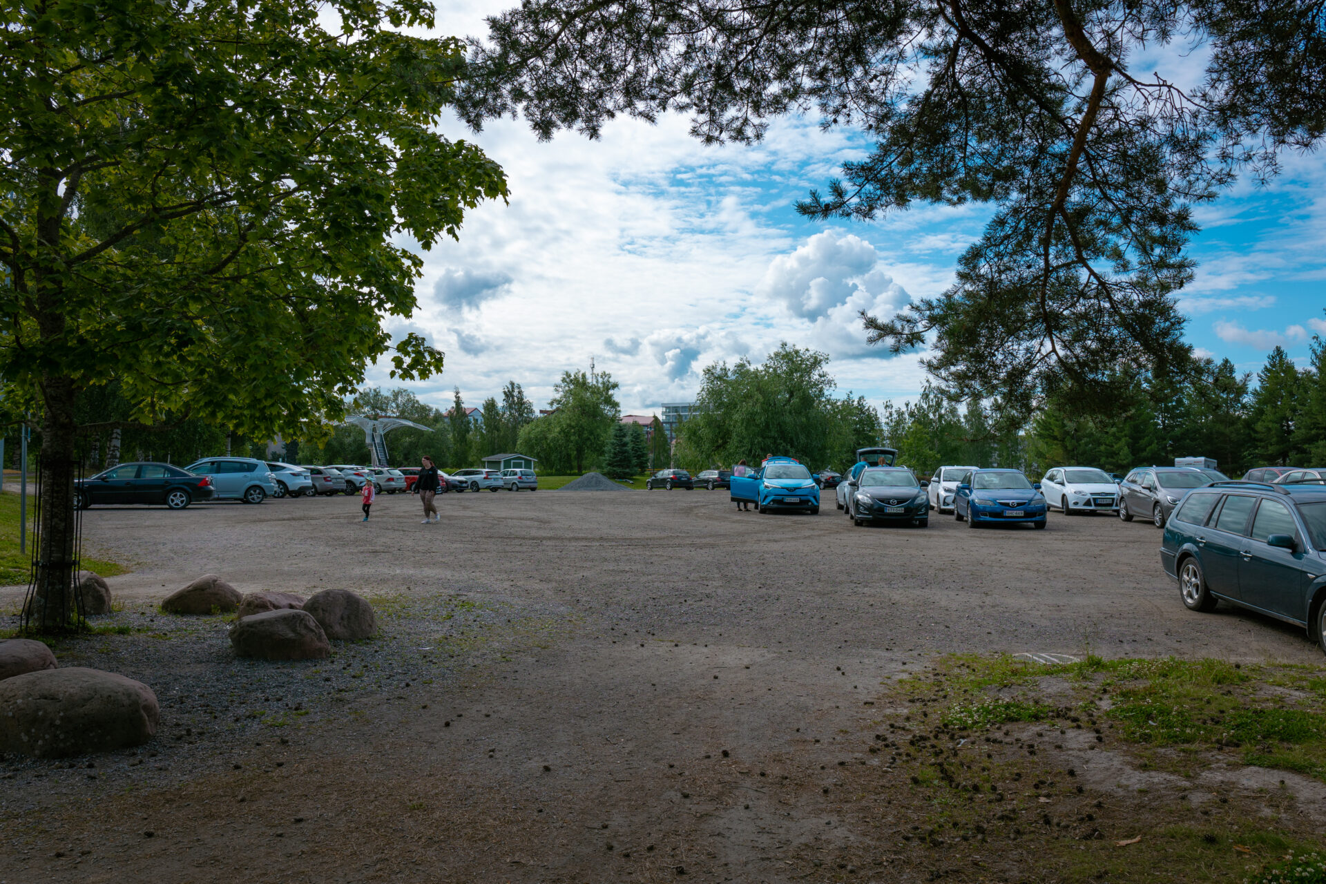 parkkipaikka jossa autoja, edustalla aikuinen ja lapsi kävelee
