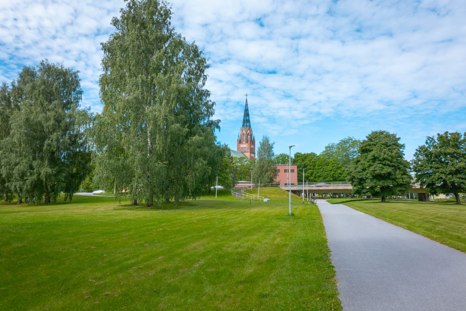 puisto jossa puita, keskellä asfalttinen pyörätie joka menee sillan alta, sillan toisella puolella kirkko
