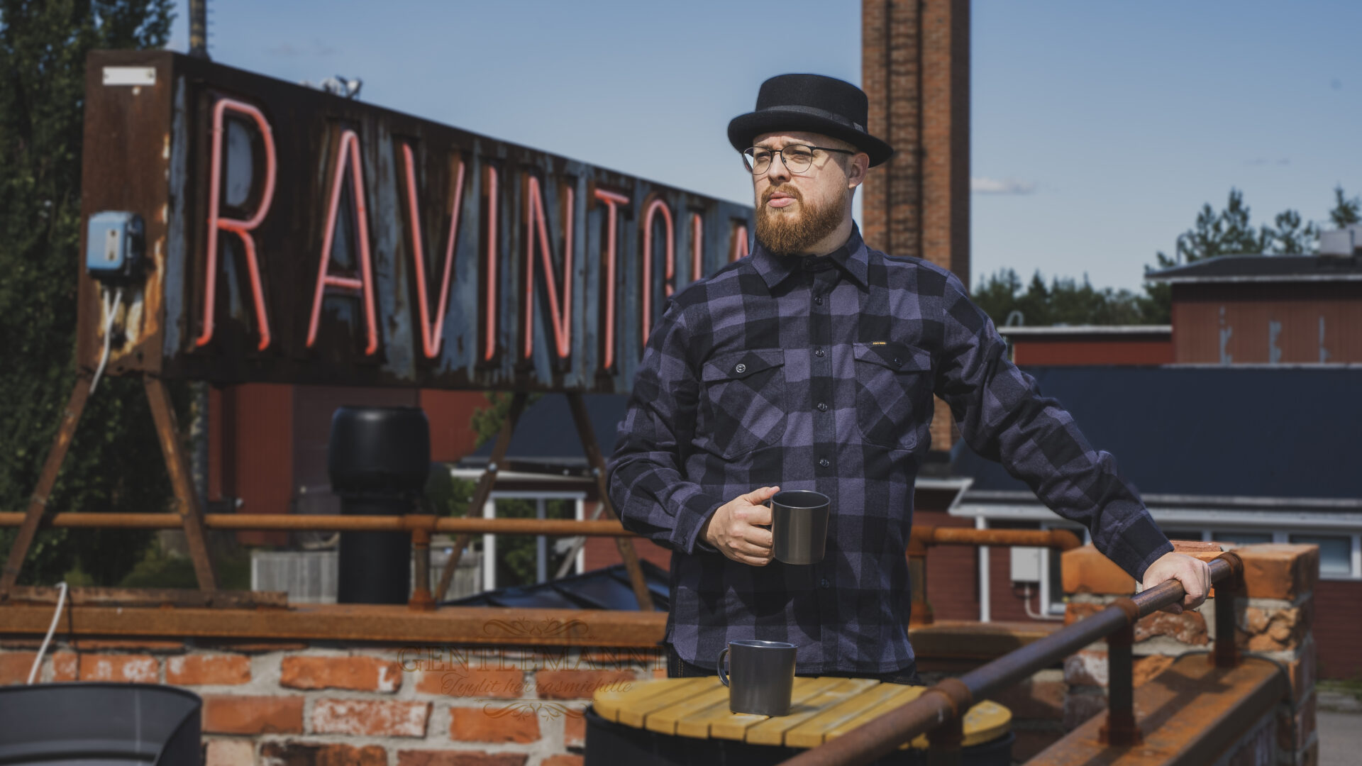 Gentlemanni: gentleman standing on the balcony with a mug in his hand wearing a checked shirt and hat.