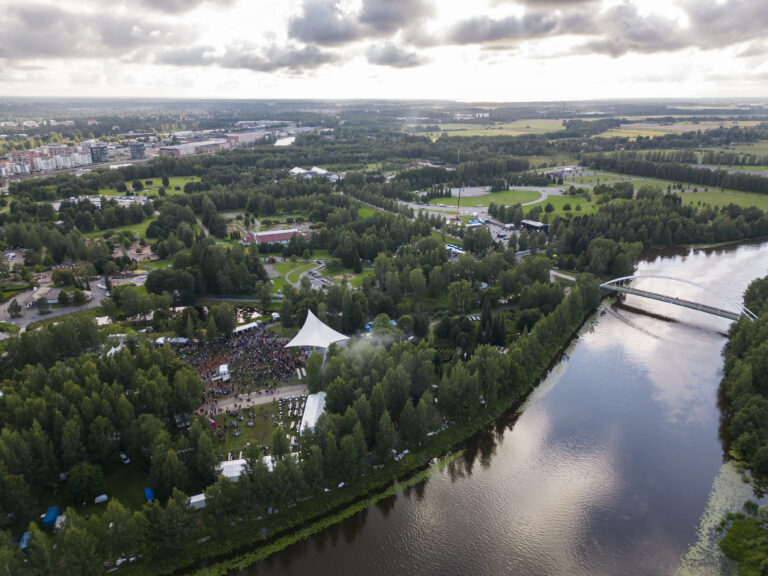 Ilmakuva vehreästä luodosta kesäisen festaritapahtuman aikaan. Featrialueen ympärillä joki ja sen ylittävä silta, peltoja, kaupunkimiljöötä ja aurinko paistaa pilvien raosta.