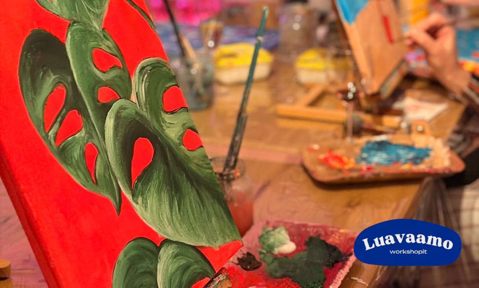 Luavaamon pöydällä valmistunut taulu, jossa punaisella pohjalla viheä peikonlehti ja taustalla henkilö maalaa omaa tauluaan.
